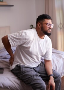 Man sitting on edge of the bed holding his lower back in pain.